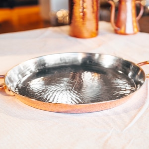 May include: A hammered copper serving tray with two handles. The tray is oval shaped and has a shiny, reflective surface.