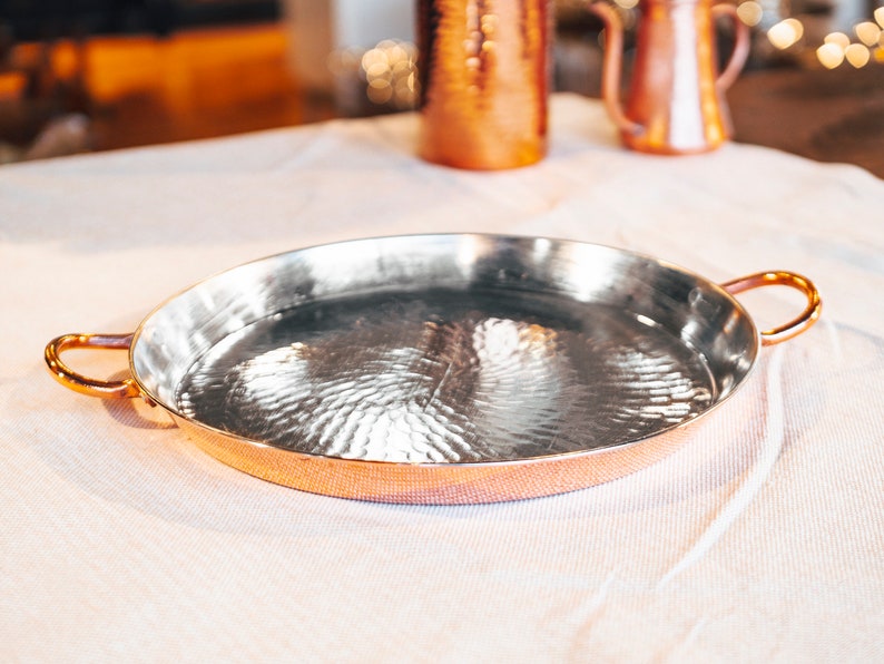 May include: A hammered copper serving tray with two handles. The tray is oval shaped and has a shiny, reflective surface.