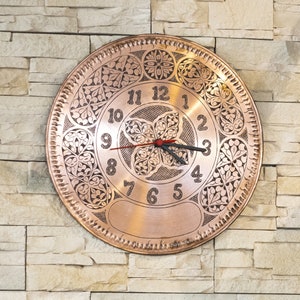 May include: A copper clock with an intricate floral design and Roman numeral markings. The clock face is surrounded by a decorative border.