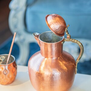 May include: A hammered copper pitcher with a lid and a brass handle. The pitcher is sitting on a white surface.