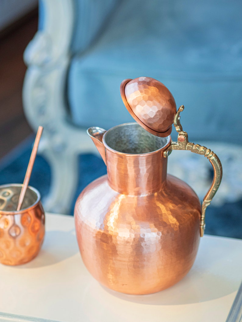 May include: A hammered copper pitcher with a lid and a brass handle. The pitcher is sitting on a white surface.