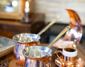 Pode incluir: Duas cafeteiras de cobre com alças de latão estão sobre uma superfície de madeira, refletindo a luz quente. Uma pequena xícara e pires de cobre também estão presentes, juntamente com uma peça de cobre em forma de meia-lua. Grãos de café espalhados por perto.
