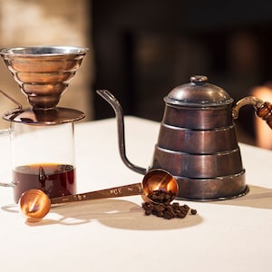 May include: Copper coffee pot with a wooden handle, a copper pour-over coffee maker with a glass carafe, a copper measuring spoon, and coffee beans on a white surface.