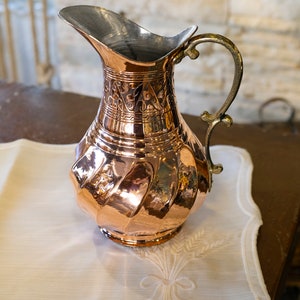May include: A copper pitcher with an ornate design and a silver handle. The pitcher is sitting on a white cloth with an embroidered design.