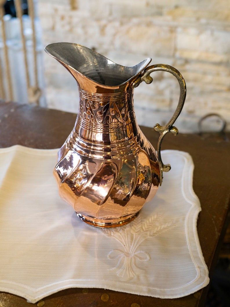 May include: A copper pitcher with an ornate design and a silver handle. The pitcher is sitting on a white cloth with an embroidered design.