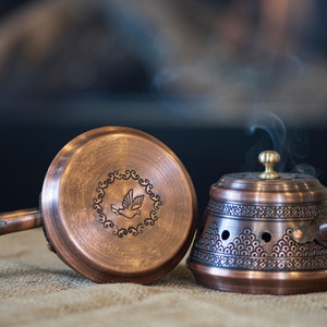 May include: Two copper incense burners with intricate designs. One burner features a bird design and the other has a geometric pattern. Both burners have black handles and are on a hessian surface. Smoke is rising from the burner with the geometric pattern.