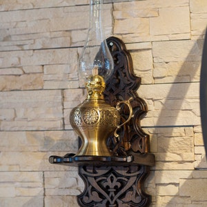 May include: A gold oil lamp with an ornate design hangs on a wooden wall mount with intricate carvings. The lamp has a clear glass chimney and a brass base.