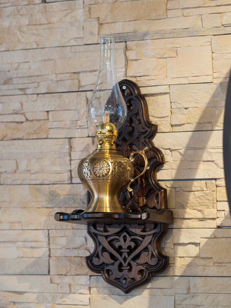 May include: A gold oil lamp with an ornate design hangs on a wooden wall mount with intricate carvings. The lamp has a clear glass chimney and a brass base.
