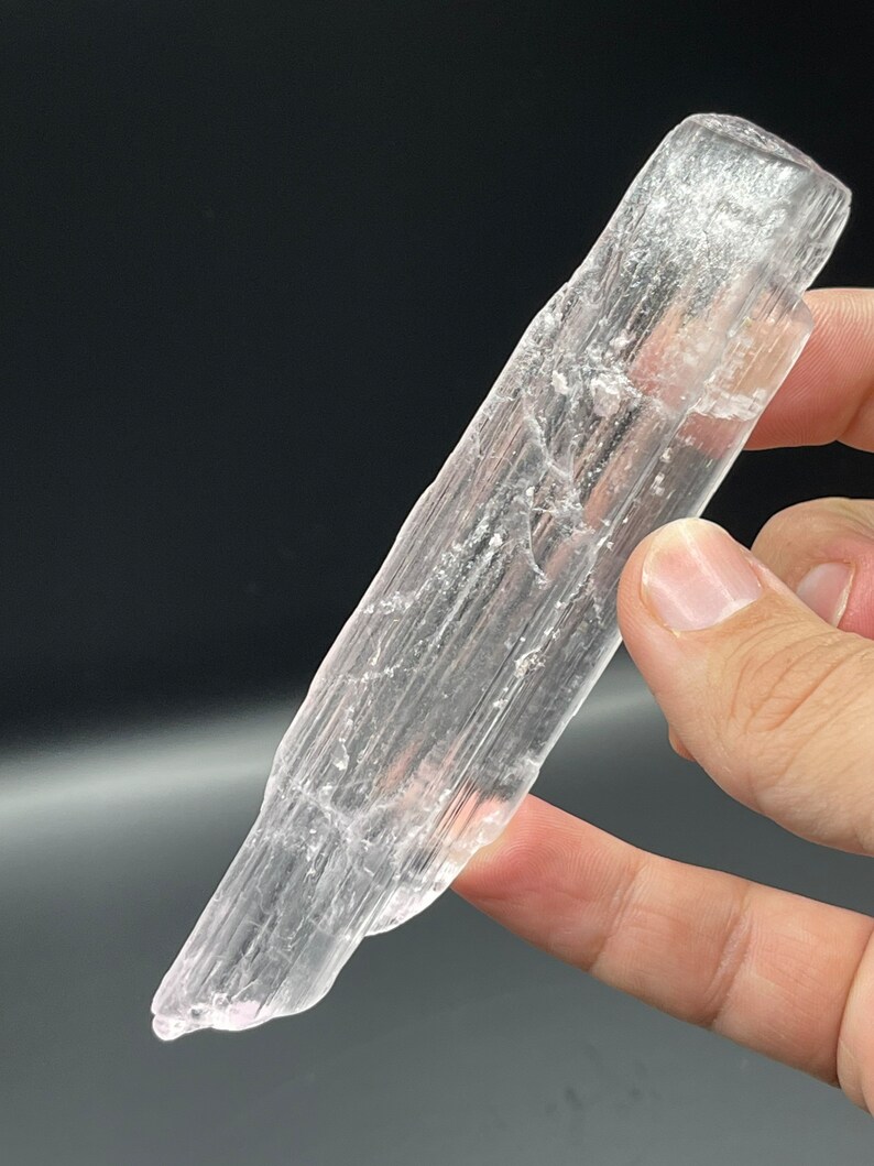 May include: A clear selenite crystal wand with a pointed tip and a smooth, polished surface. The crystal is translucent and has a slight pink hue.