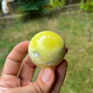 May include: A smooth, yellow, polished sphere of yellow calcite, a type of crystal, is held in a hand.