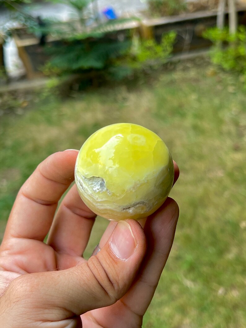 May include: A smooth, yellow, polished sphere of yellow calcite, a type of crystal, is held in a hand.