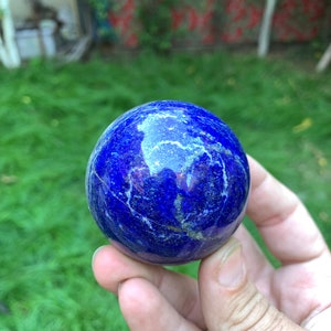 May include: A smooth, polished, blue lapis lazuli sphere with white veins and gold flecks. The sphere is being held in a hand.