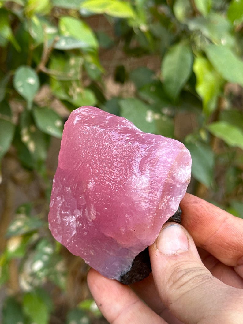 May include: A large, pink, translucent crystal with a rough, natural surface. The crystal is being held in a hand, and the background is blurred.