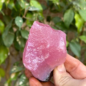 May include: A large, pink, rough-cut crystal with a smooth, polished surface. The crystal has a light pink hue with a slight purple tint.