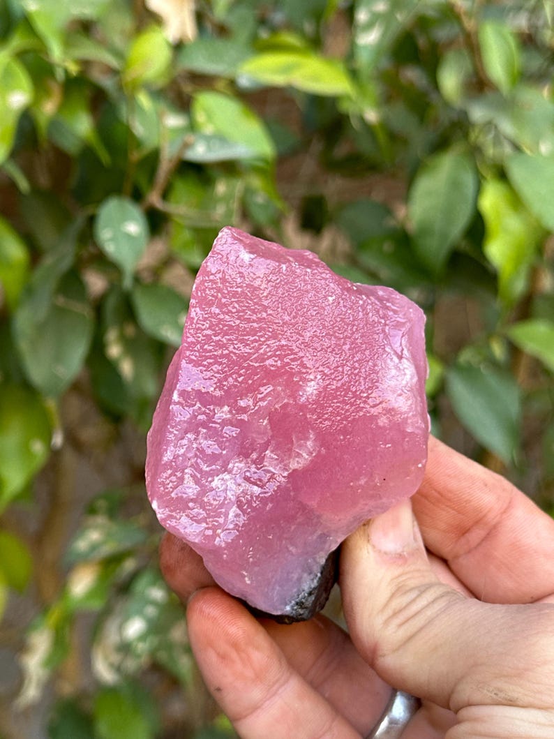 May include: A large, pink, rough-cut crystal with a smooth, polished surface. The crystal has a light pink hue with a slight purple tint.