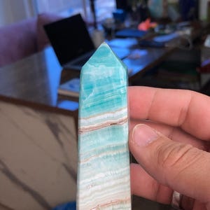 May include: A blue and white banded aquamarine crystal point, also known as a tower or obelisk, is held in a hand. The crystal is polished and has a smooth, glassy surface.