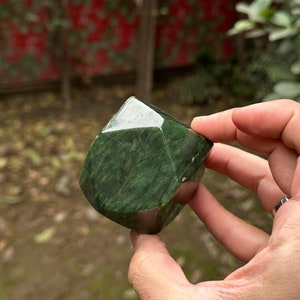 May include: A dark green, faceted, polished stone with a marbled pattern. The stone is held in a hand.
