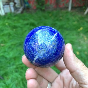 May include: A smooth, blue lapis lazuli sphere with white veins and gold flecks. The sphere is being held in a hand.