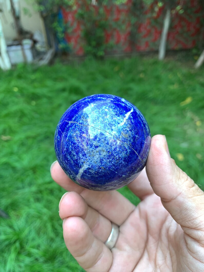 May include: A smooth, blue lapis lazuli sphere with white veins and gold flecks. The sphere is being held in a hand.