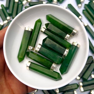 Op de afbeelding: Een verzameling groene jade stenen hangers met zilveren dopjes. De hangers zijn in de vorm van lange, dunne rechthoeken met puntige uiteinden. Ze zijn gerangschikt in een witte kom.