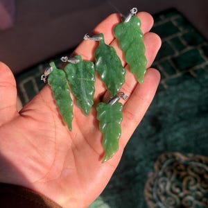Puede incluir: Una mano sostiene seis colgantes de hojas de jade verde con aros plateados. Los colgantes están tallados con detalles de venas de hojas y tienen un acabado pulido y liso. El fondo es una superficie verde estampada.