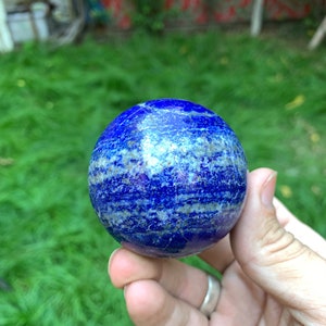 May include: A smooth, polished blue lapis lazuli sphere with white and gold veins. The sphere is being held in a hand.