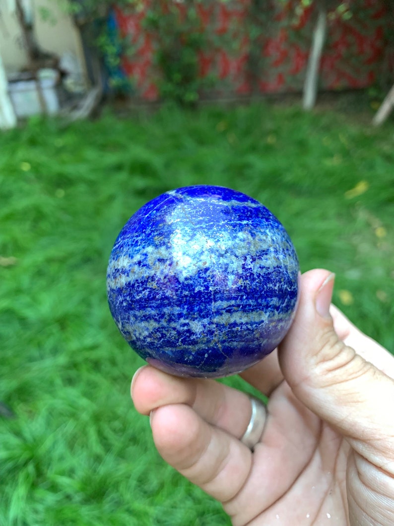 May include: A smooth, polished blue lapis lazuli sphere with white and gold veins. The sphere is being held in a hand.
