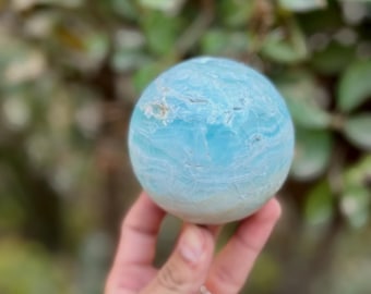 May include: A light blue sphere with white and light blue bands. The sphere is held in a hand, with a blurred green background. The sphere is a decorative item, possibly a crystal or stone.