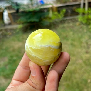 May include: A yellow and white polished sphere of yellow calcite, a type of crystal, is held in a hand.