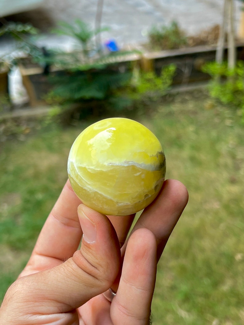 May include: A yellow and white polished sphere of yellow calcite, a type of crystal, is held in a hand.