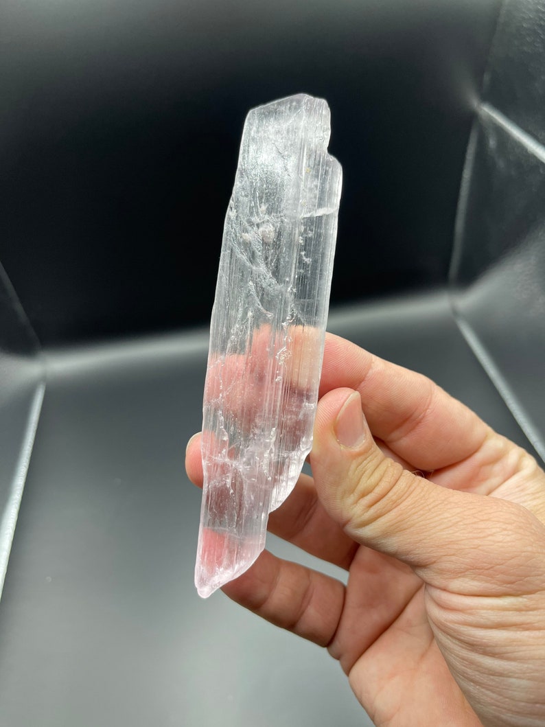 May include: A clear, translucent selenite crystal with a pointed tip. The crystal has a natural, rough texture and is approximately 5 inches long.