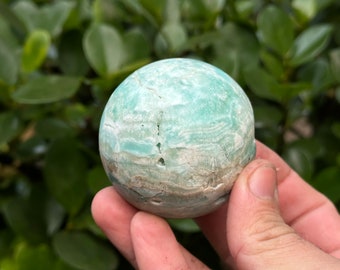 May include: A green and white sphere of polished selenite crystal. The sphere has a smooth, polished surface and is a light green color with white streaks.