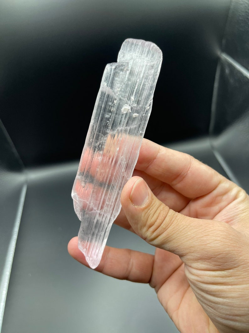 May include: A clear selenite crystal with a striated texture. The crystal is about 5 inches long and has a slightly pink hue.