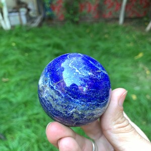 May include: A smooth, blue lapis lazuli sphere with white veins. The sphere is being held in a hand.
