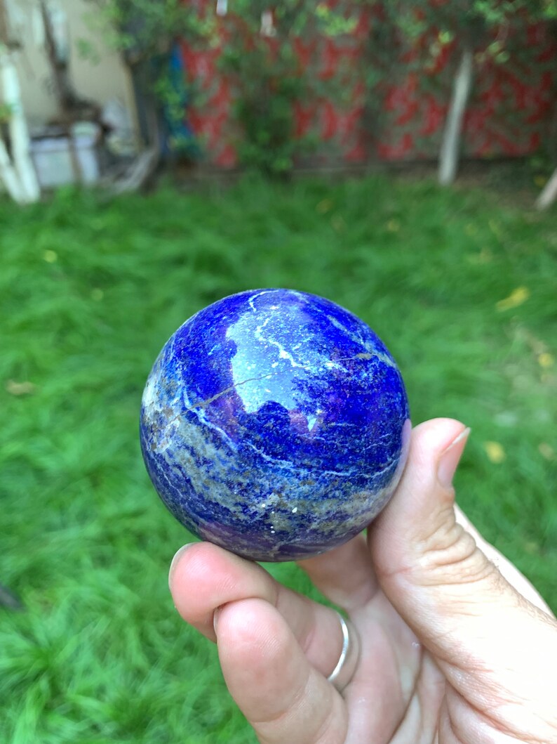 May include: A smooth, blue lapis lazuli sphere with white veins. The sphere is being held in a hand.