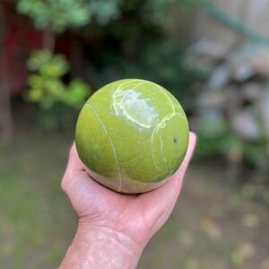 Könnte beinhalten: Eine grüne Kugel mit weißen Adern, poliert zu einem glatten Finish. Die Kugel wird in einer Hand gehalten.