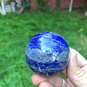 May include: A smooth, polished blue lapis lazuli sphere with white and gold veins. The sphere is held in a hand.