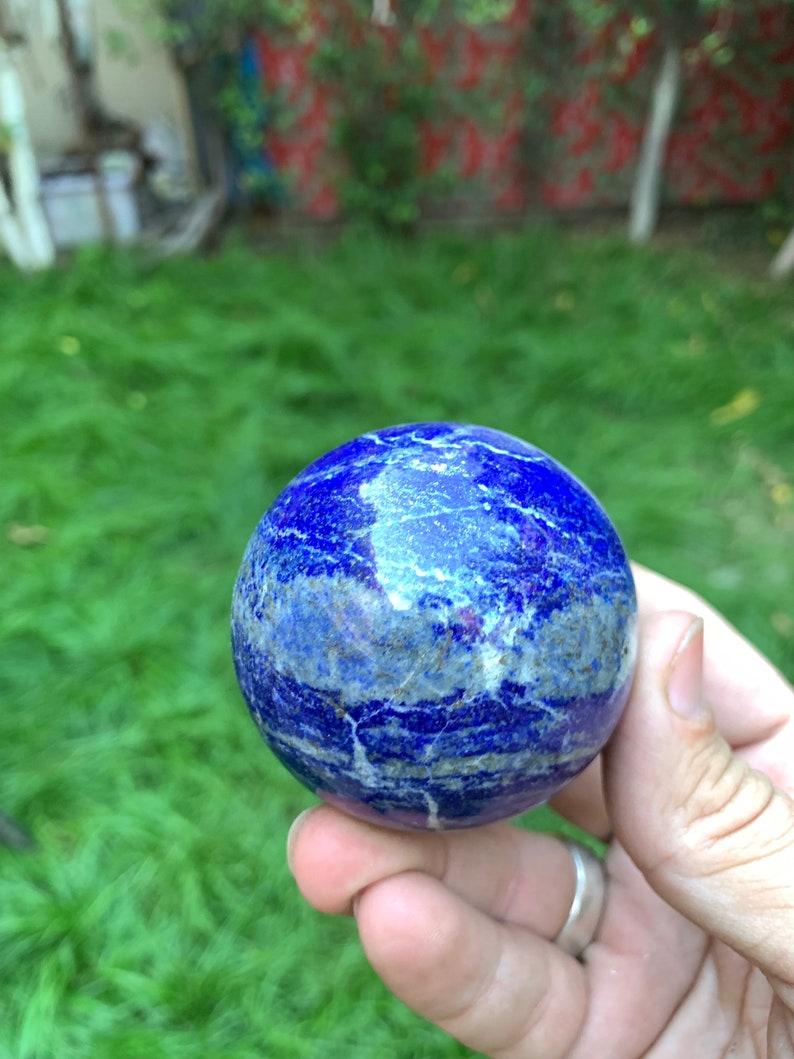 May include: A smooth, polished blue lapis lazuli sphere with white and gold veins. The sphere is held in a hand.