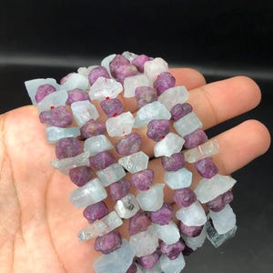 以下が含まれることがあります： 薄い青色のアクアマリンと濃い紫色のルビーのビーズが交互に並んだ宝石ビーズのストランド。ビーズはファセットカットされており、粗くて自然な外観をしています。