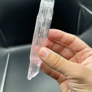 May include: A clear selenite crystal wand with a pink hue. The wand is smooth and has a pointed tip.