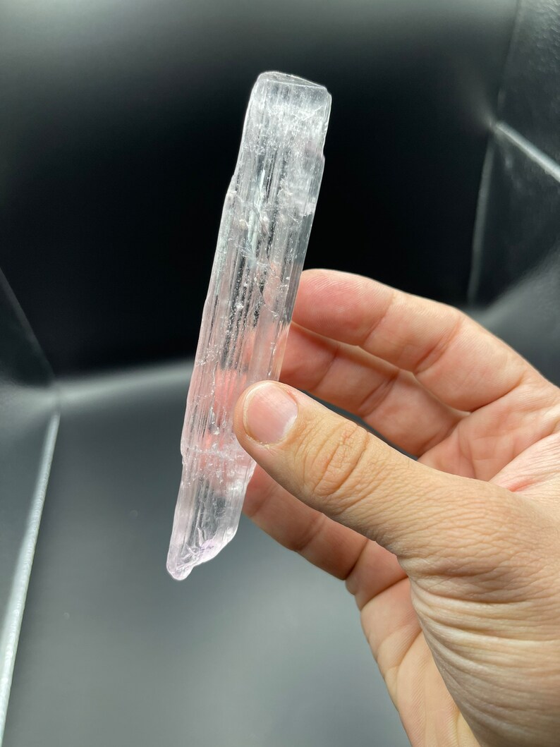 May include: A clear selenite crystal wand with a pink hue. The wand is smooth and has a pointed tip.