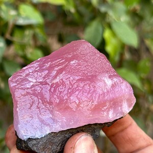 May include: A large, pink, translucent, rough-cut gemstone with a dark gray base. The gemstone has a smooth, polished surface and is a natural, raw crystal.