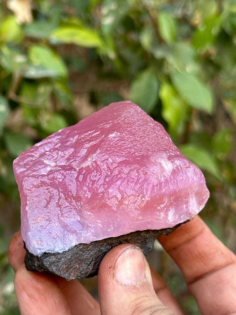 May include: A large, pink, translucent, rough-cut gemstone with a dark gray base. The gemstone has a smooth, polished surface and is a natural, raw crystal.