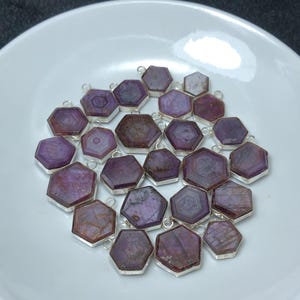 May include: A collection of hexagonal, purple gemstone pendants with silver-colored metal edges. The gemstones have a mix of light and dark purple hues. The pendants are arranged on a white plate, creating a visually appealing display.