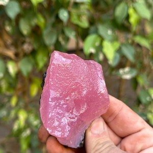 May include: A large, pink, translucent crystal with a rough, irregular surface. The crystal is held in a hand, and the background is blurred.