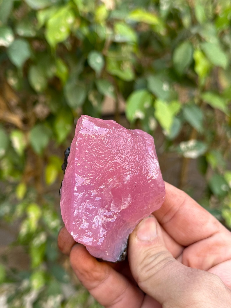 May include: A large, pink, translucent crystal with a rough, irregular surface. The crystal is held in a hand, and the background is blurred.