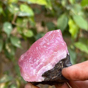May include: A pink and white crystal with a rough, natural texture. The crystal is attached to a dark gray rock.