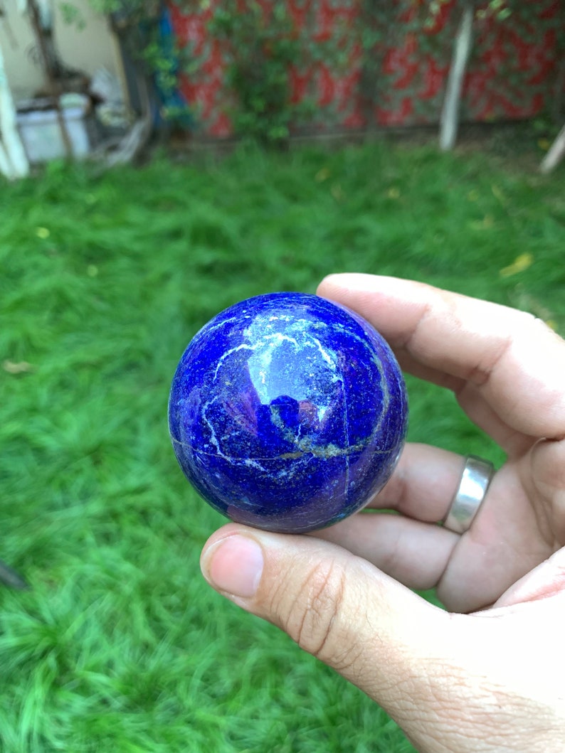 May include: A smooth, round, blue lapis lazuli sphere with white veins. The sphere is being held in a hand.
