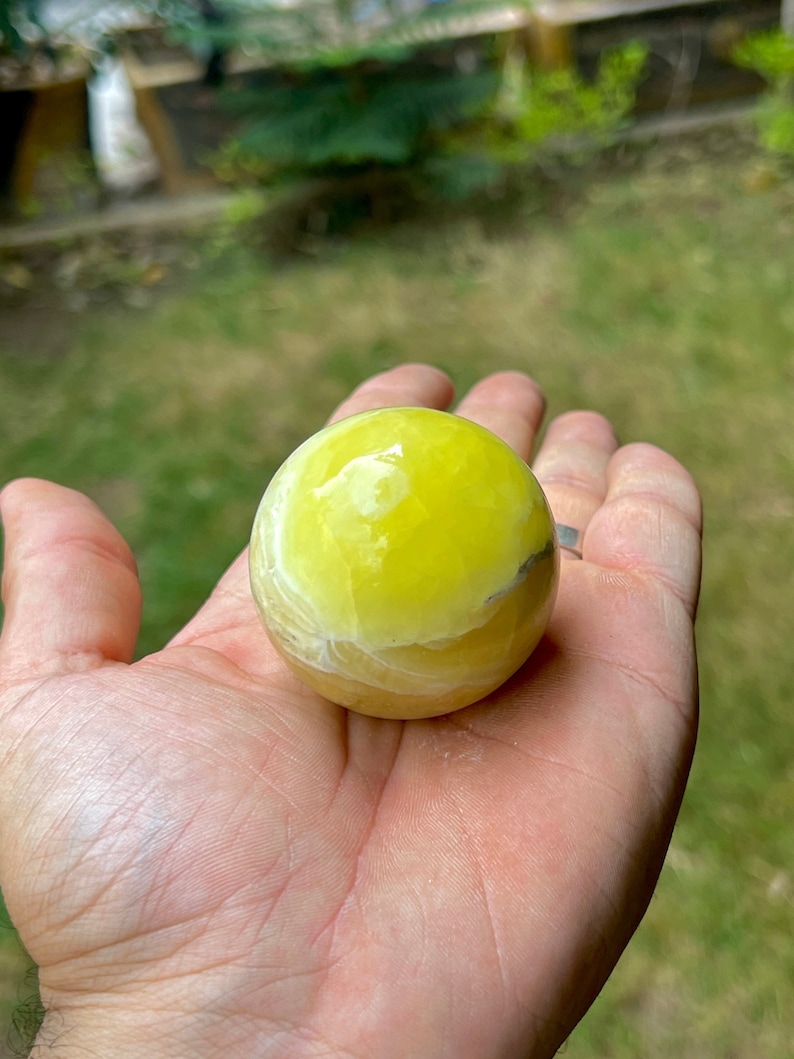 May include: A yellow and white polished sphere of yellow calcite, a type of crystal, is held in a person's hand.