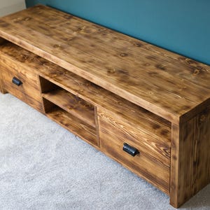 May include: A wooden TV stand with a warm brown finish. The stand features two drawers with black handles and open shelving. The wood grain is visible, adding texture. The stand is placed on a light gray carpet, with a teal wall in the background.